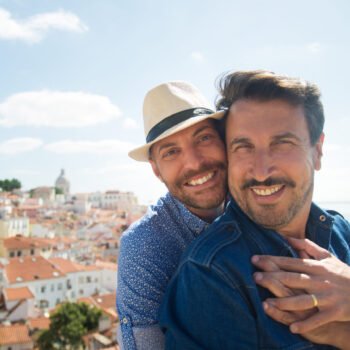 Loving gay couple taking selfie on balcony. Two men in casual clothes smiling at camera, city in background. Love, affection, relationship, travelling concept